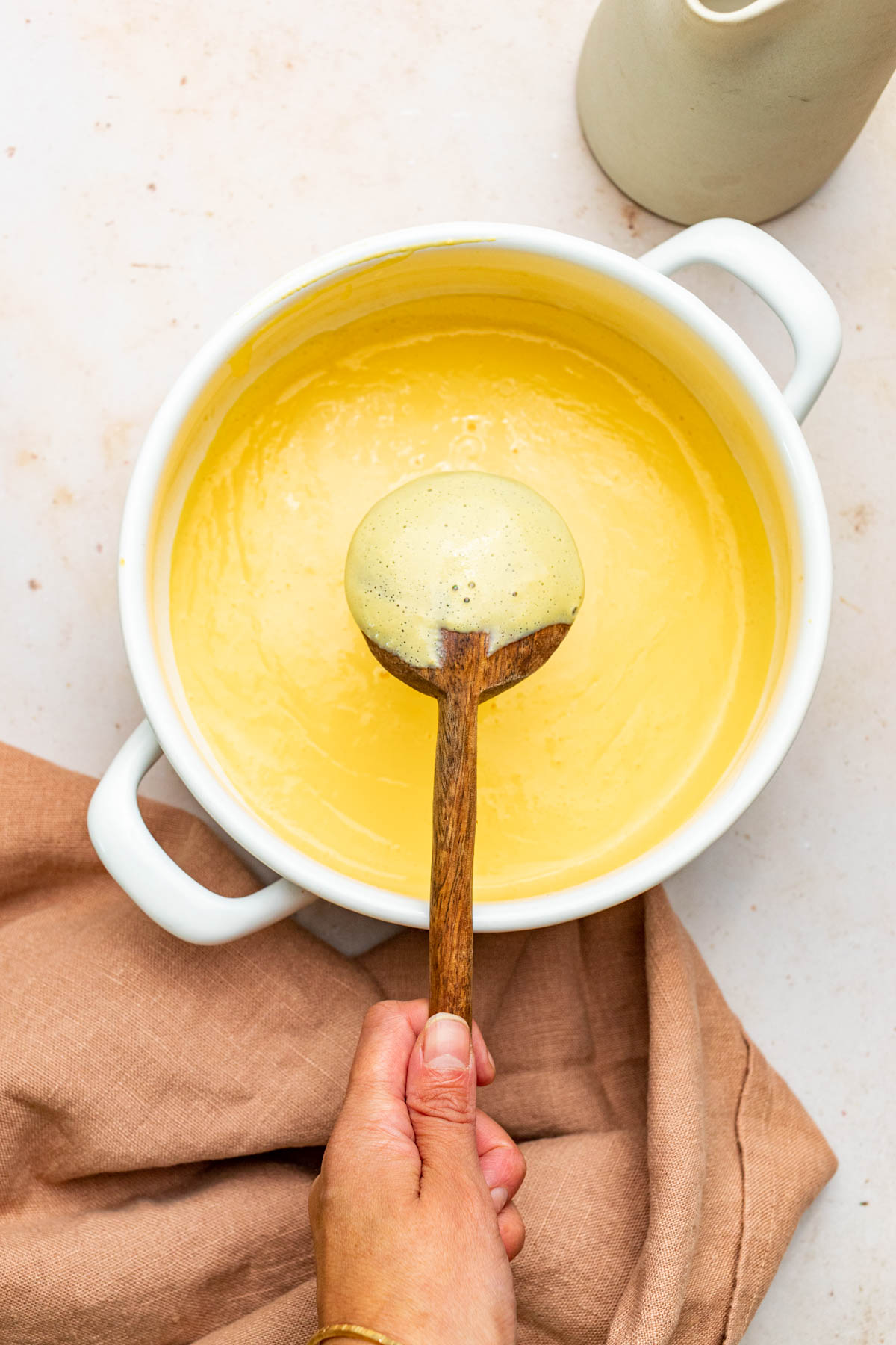 A hand holding a wooden spoon over a pot of custard to show the thickness of the custard.