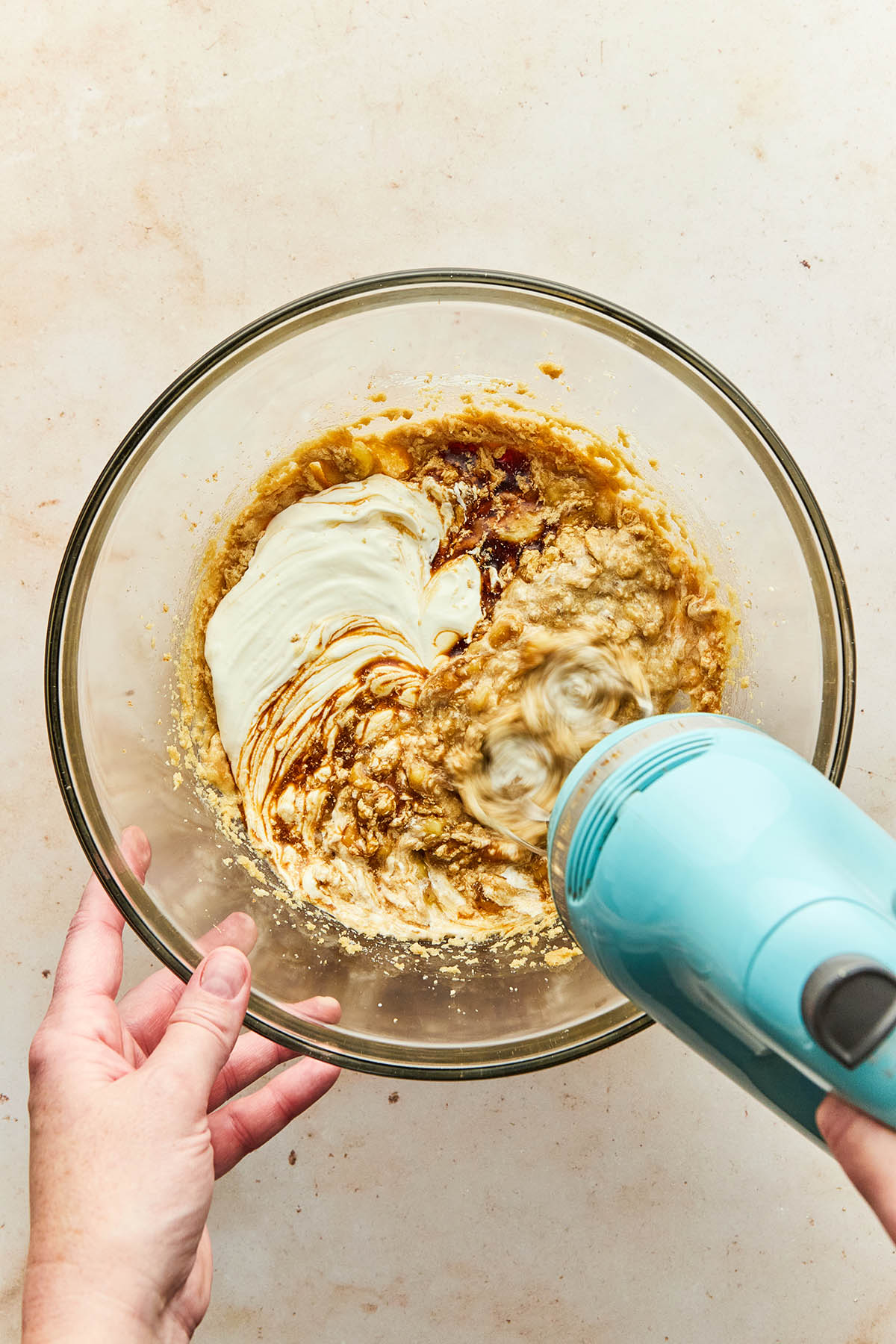 A hand using a hand mixer to mix wet ingredients in a glass bowl.