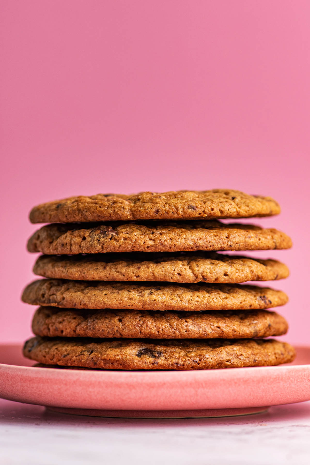 Stack of thin chocolate chunk cookies on a pink plate in front of a pink background.