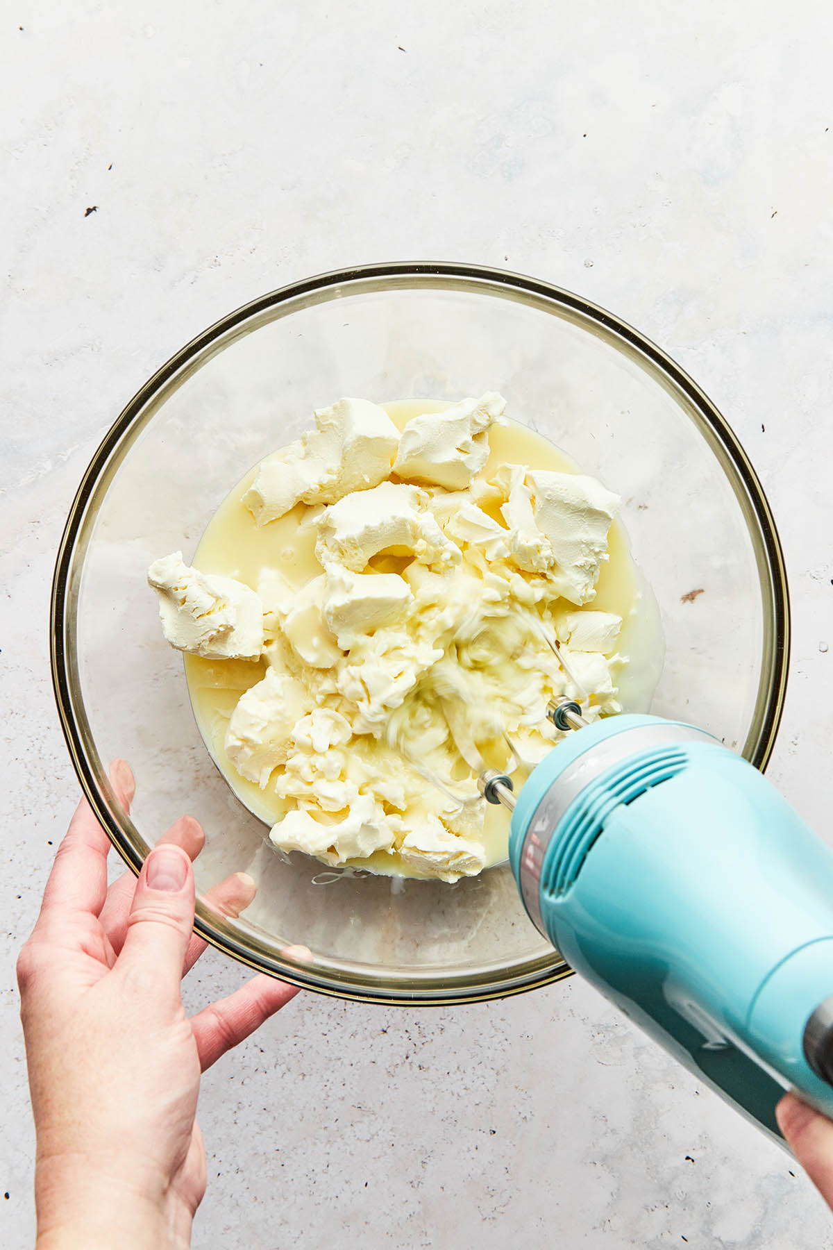A hand using a handheld mixer to beat cream cheese and sweetened condensed milk in a bowl.
