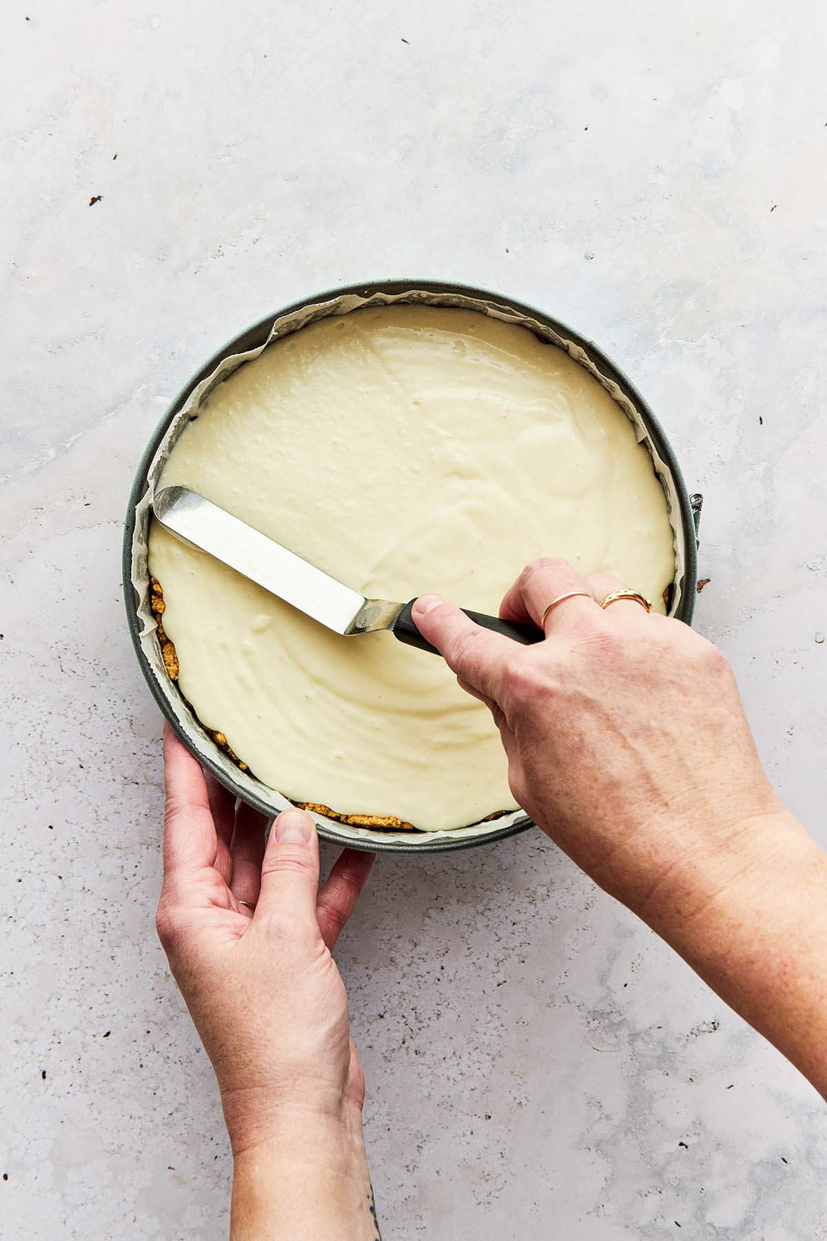 A hand using a small offset aptula to smooth cheesecake filling in a tin.