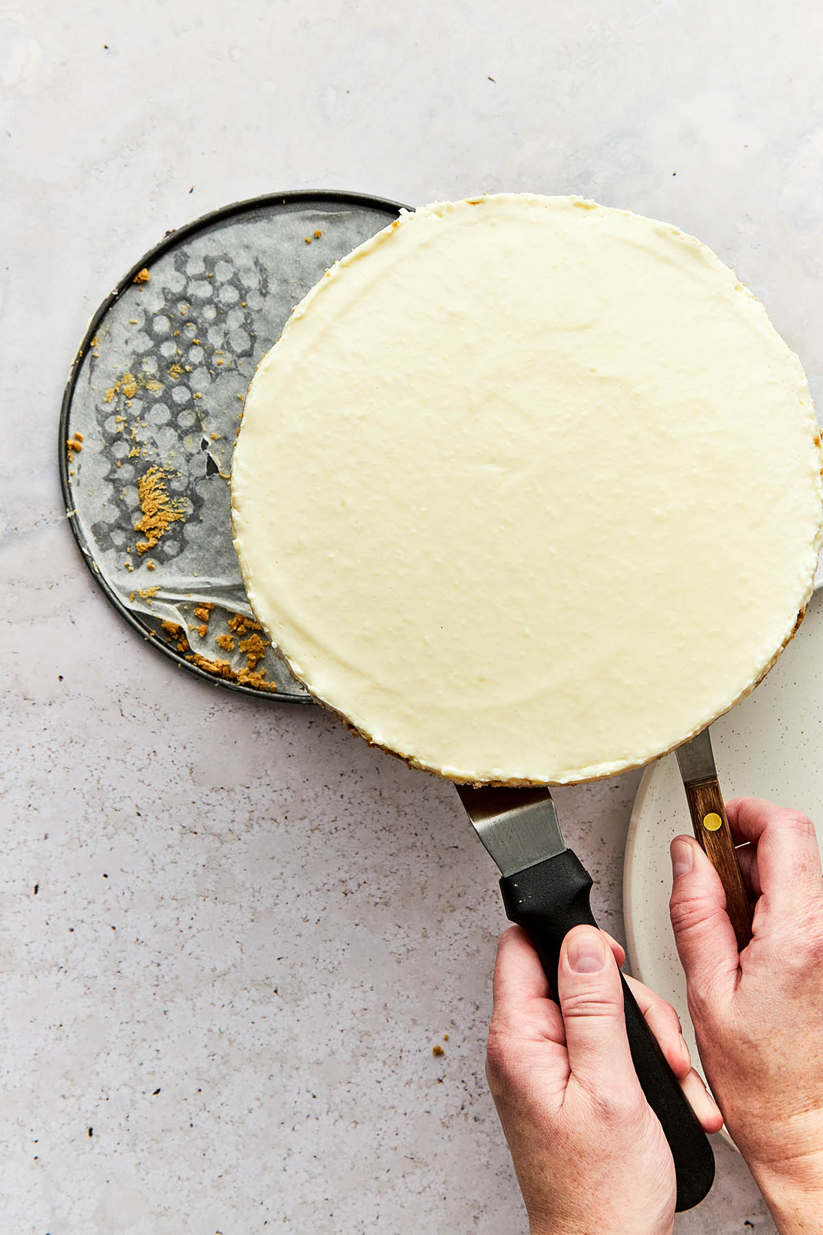 Hands using two cake spatulas to lift a whole cheesecake from the bottom of a springform pan.
