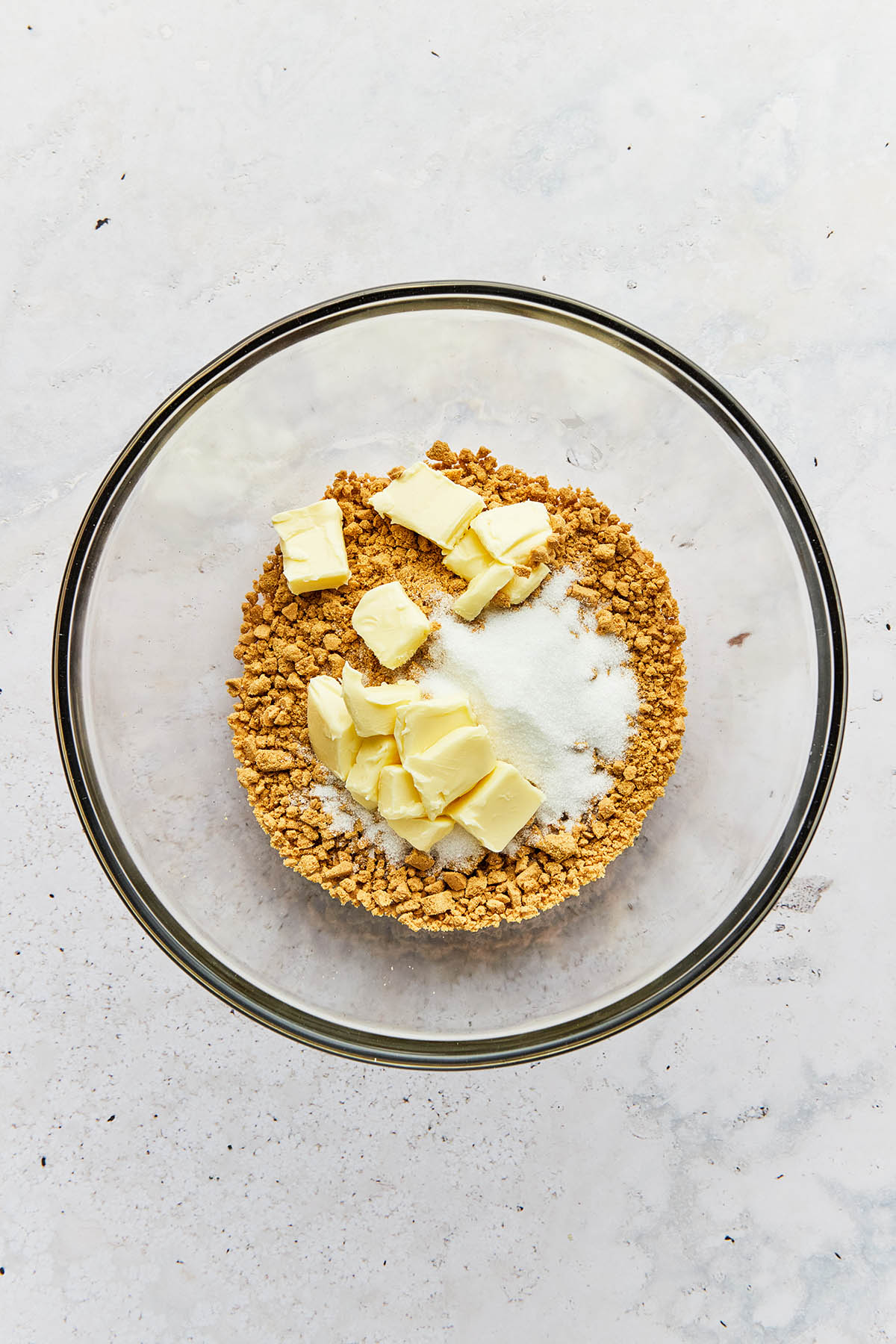 Crushed gluten-free graham cracker crumbs in a mixing bowl with chunks of butter and granulated sugar.