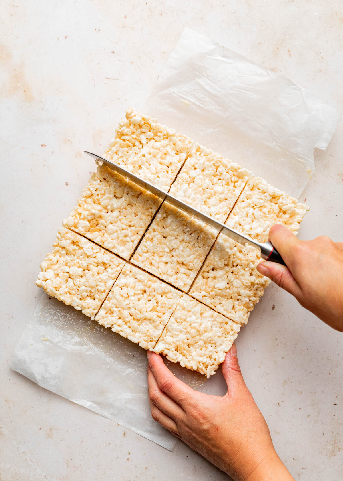 A hand slicing puffed rice marshmallow treats into nine squares with a long knife.