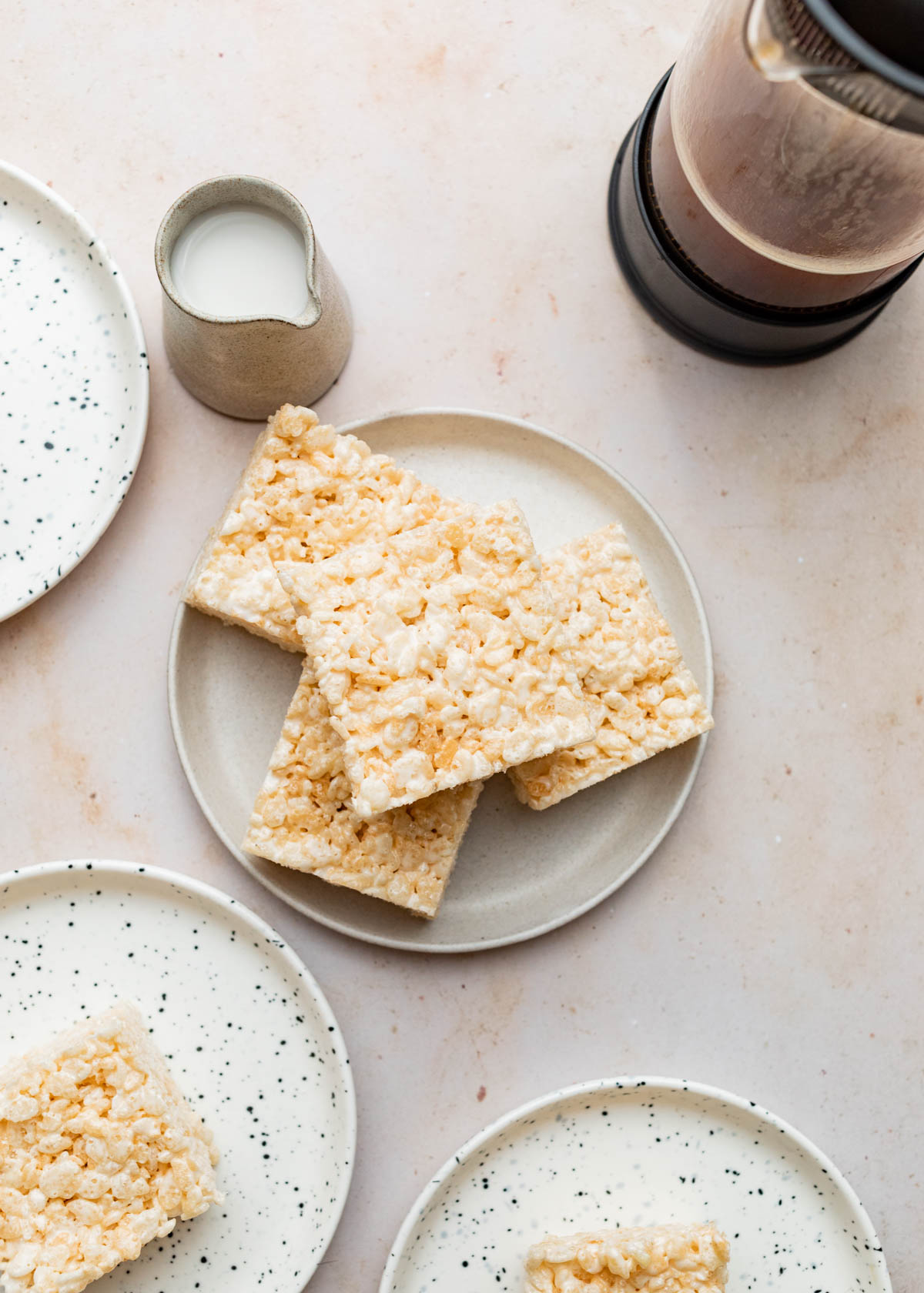 A stack of four Rice Krispie treats on a small plate with three other plates, a small pitcher of milk, and a carafe of coffee nearby.