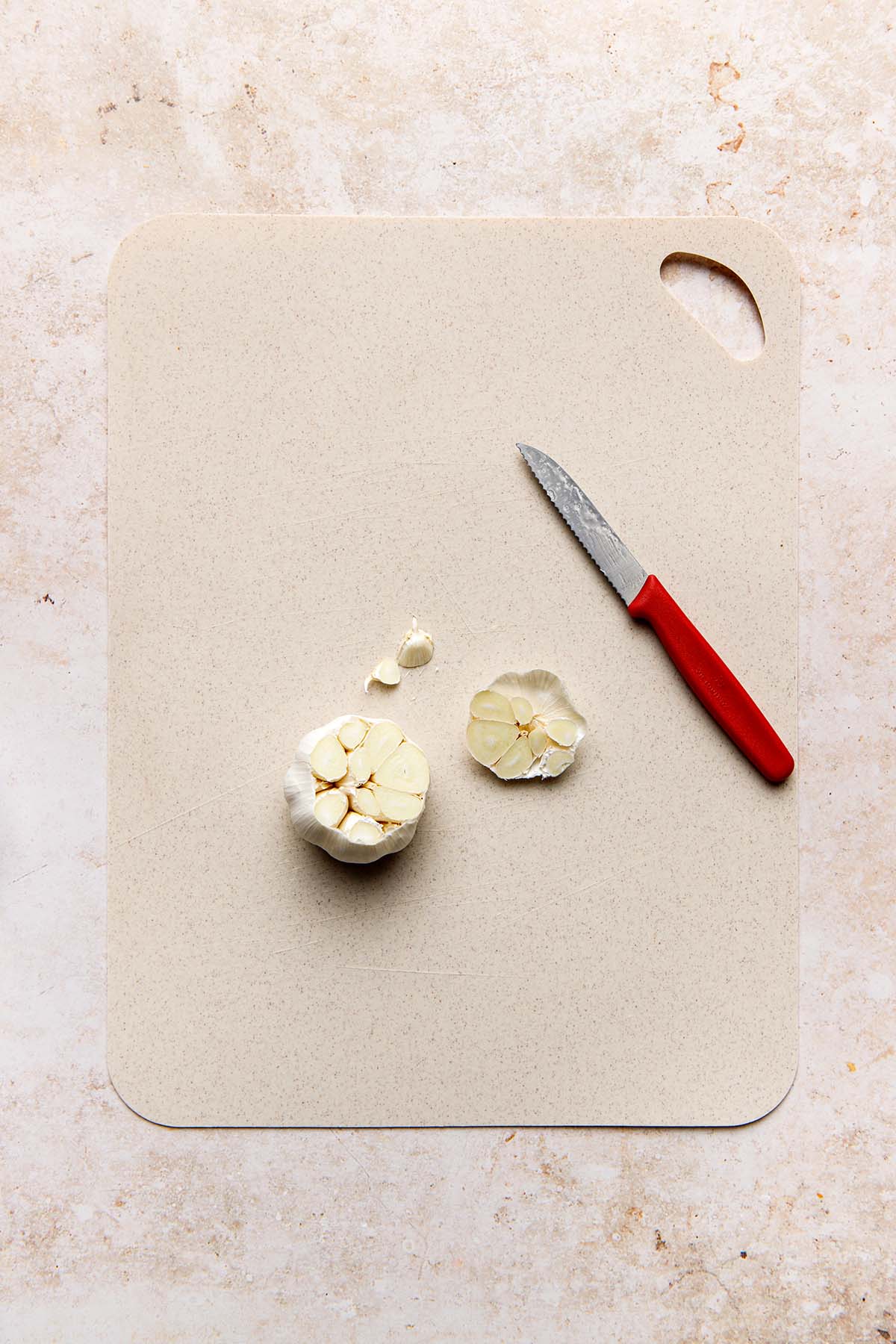 A bulb of garlic on a cutting board with the top sliced off and a small red-handled knife nearby.