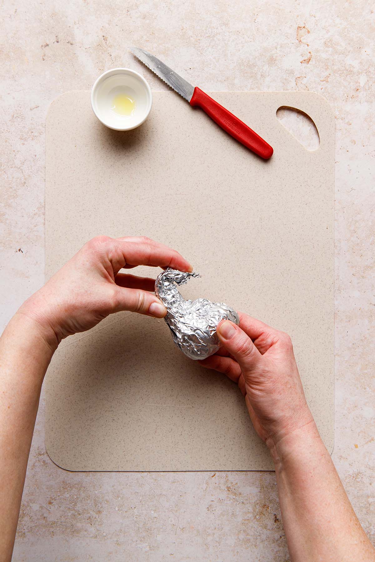 Hands wrapping a whole bulb of garlic in foil.