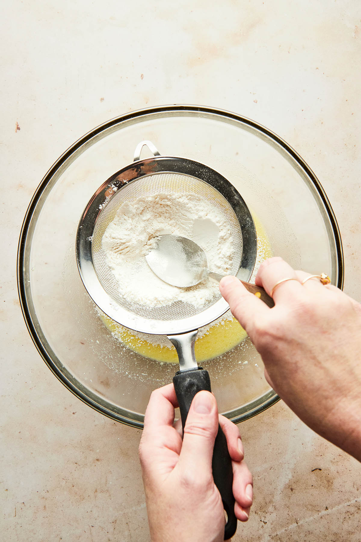 A hand using the back of a soup spoon to press flour through a sieve into a bowl of wet ingredients.