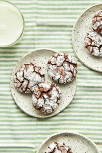 Three plates of chocolate peppermint crinkle cookies on a green and white striped cloth with a glass of milk nearby.