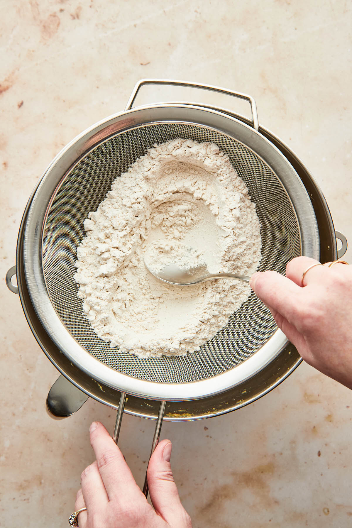 A hand using the back of a spoon to press flour through a fine mesh sieve.