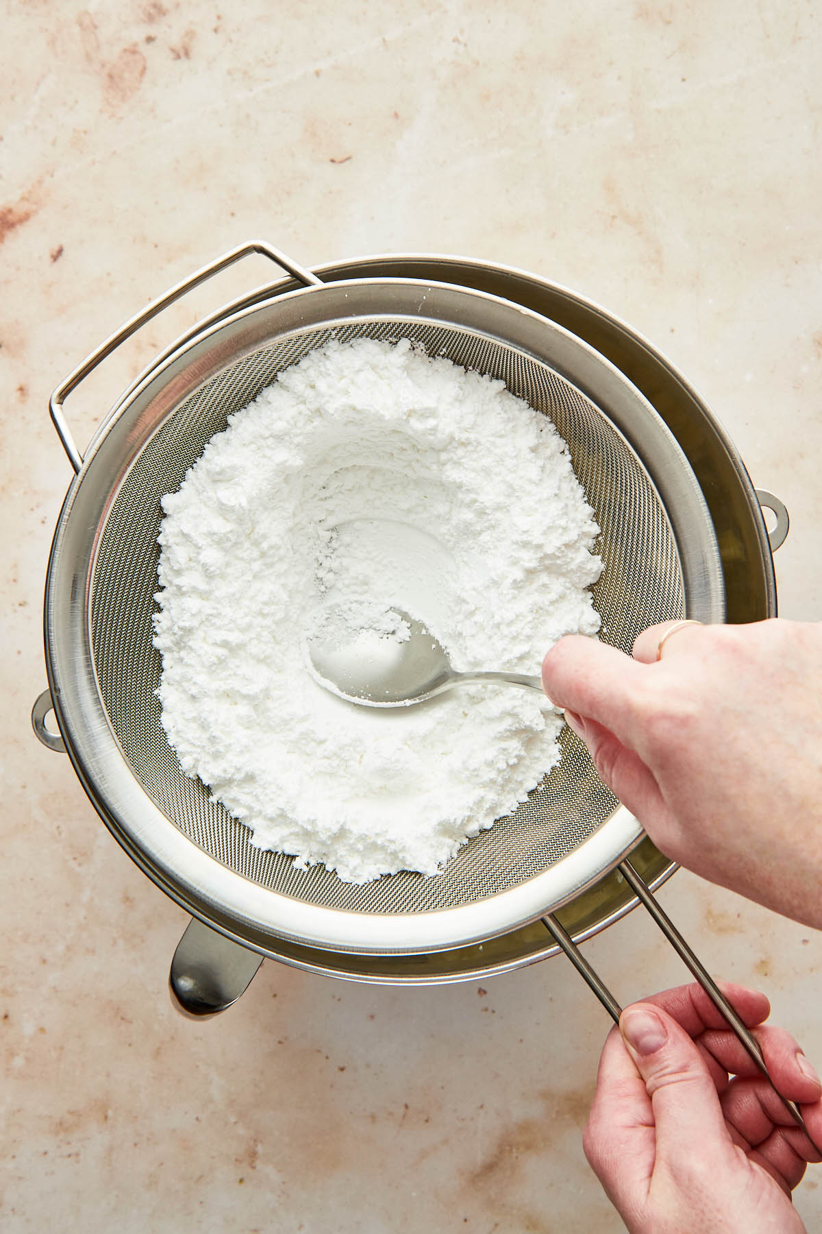 A hand using the back of a spoon to press powdered sugar through a large fine mesh sieve into a mixing bowl.