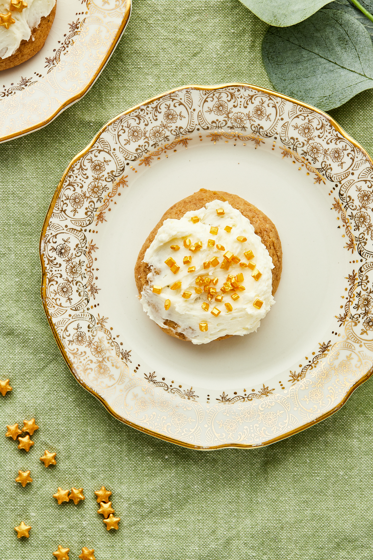 A frosted eggnog cookie with gold sprinkles on a gold rimmed plate on a green tablecloth.