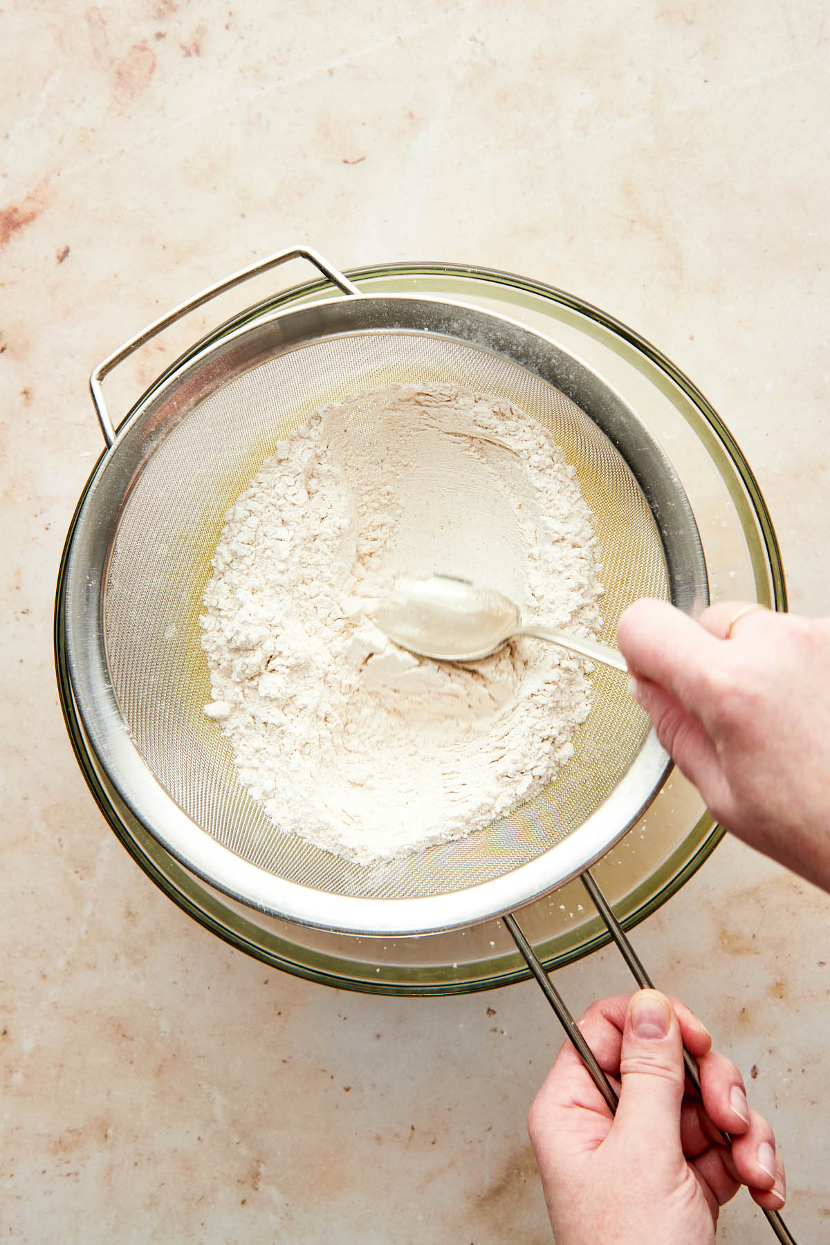 A hand using the back of a spoon to press flour through a sieve into a mixing bowl.