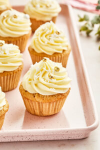 Close up image of eggnog cupcakes with eggnog buttercream frosting on a light pink baking sheet with greenery and pink fabric tucked into the top right corner.