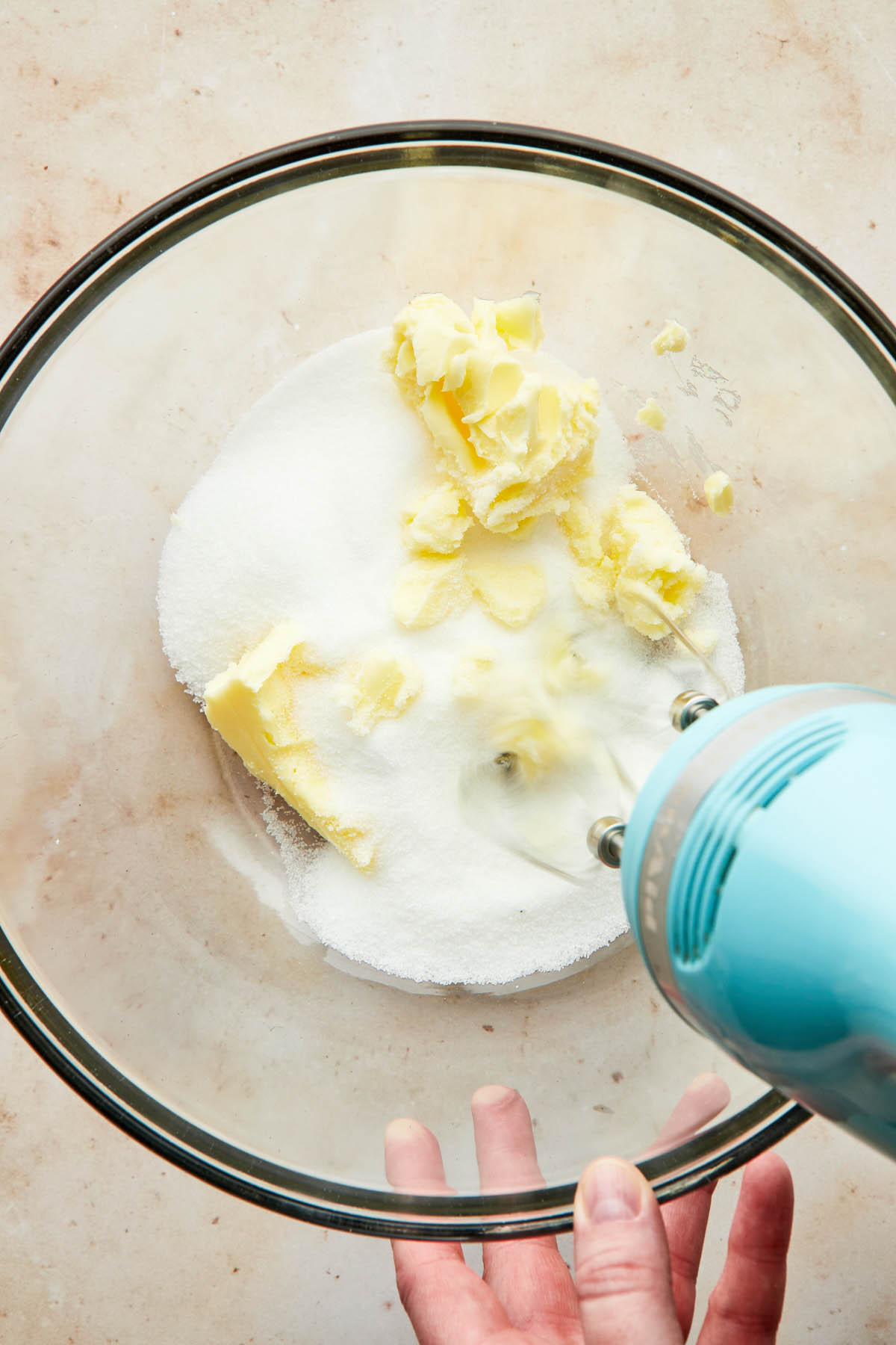 A hand using a hand moxer to cream butter and sugar together in a glass mixing bowl.