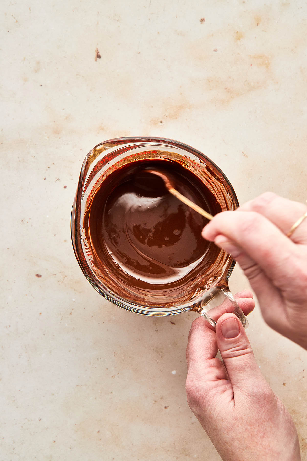 A hand stirring melted chocolate in a glass measuring cup.