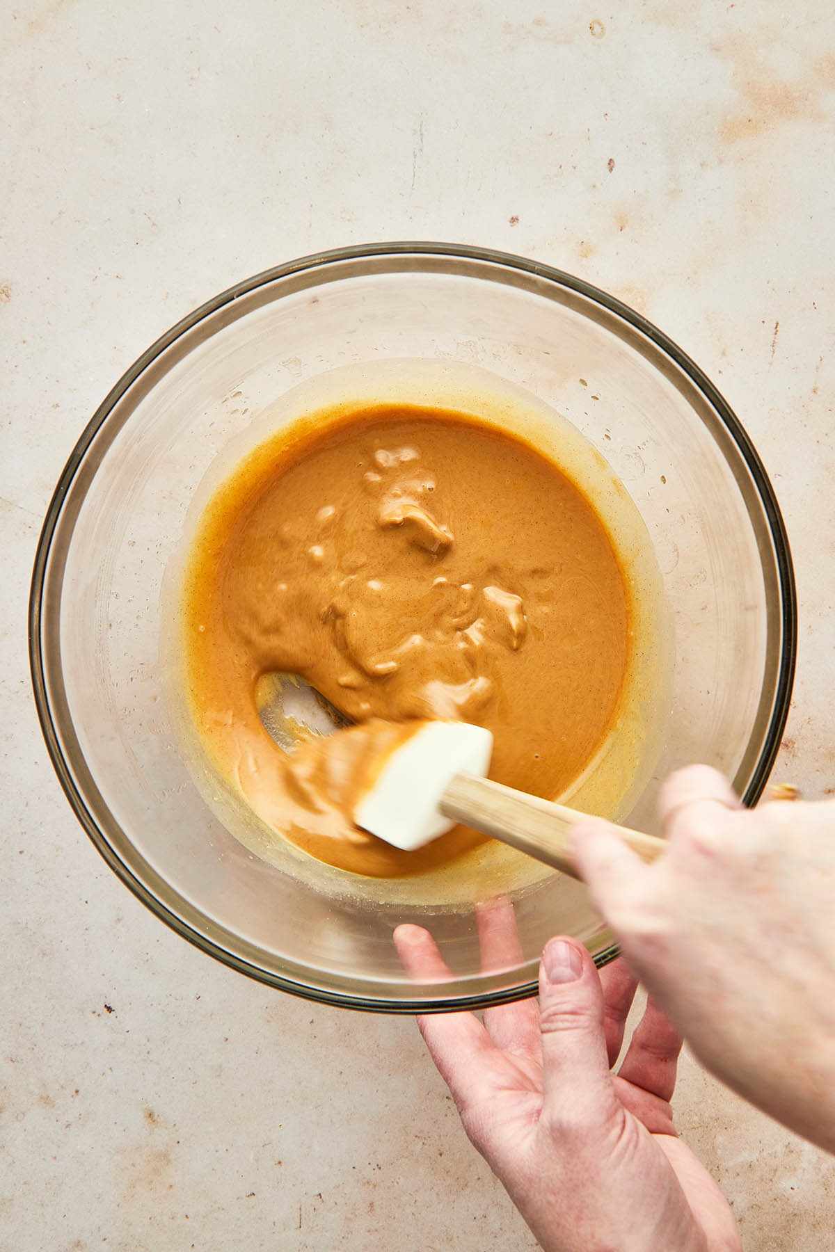 A hand stirring peanut butter in a glass micing bowl using a rubber spatula.