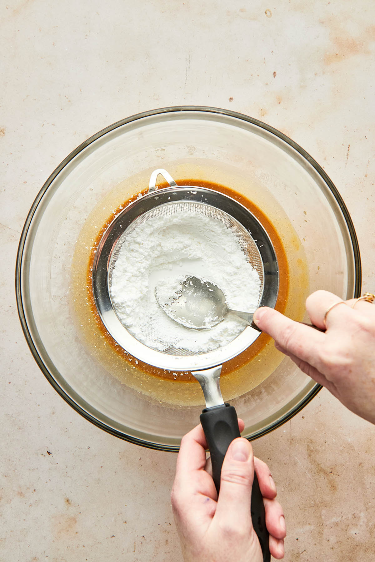 A hand using a spoon to sift powdered sugar into a glass mixing bowl with peanut butter inside.