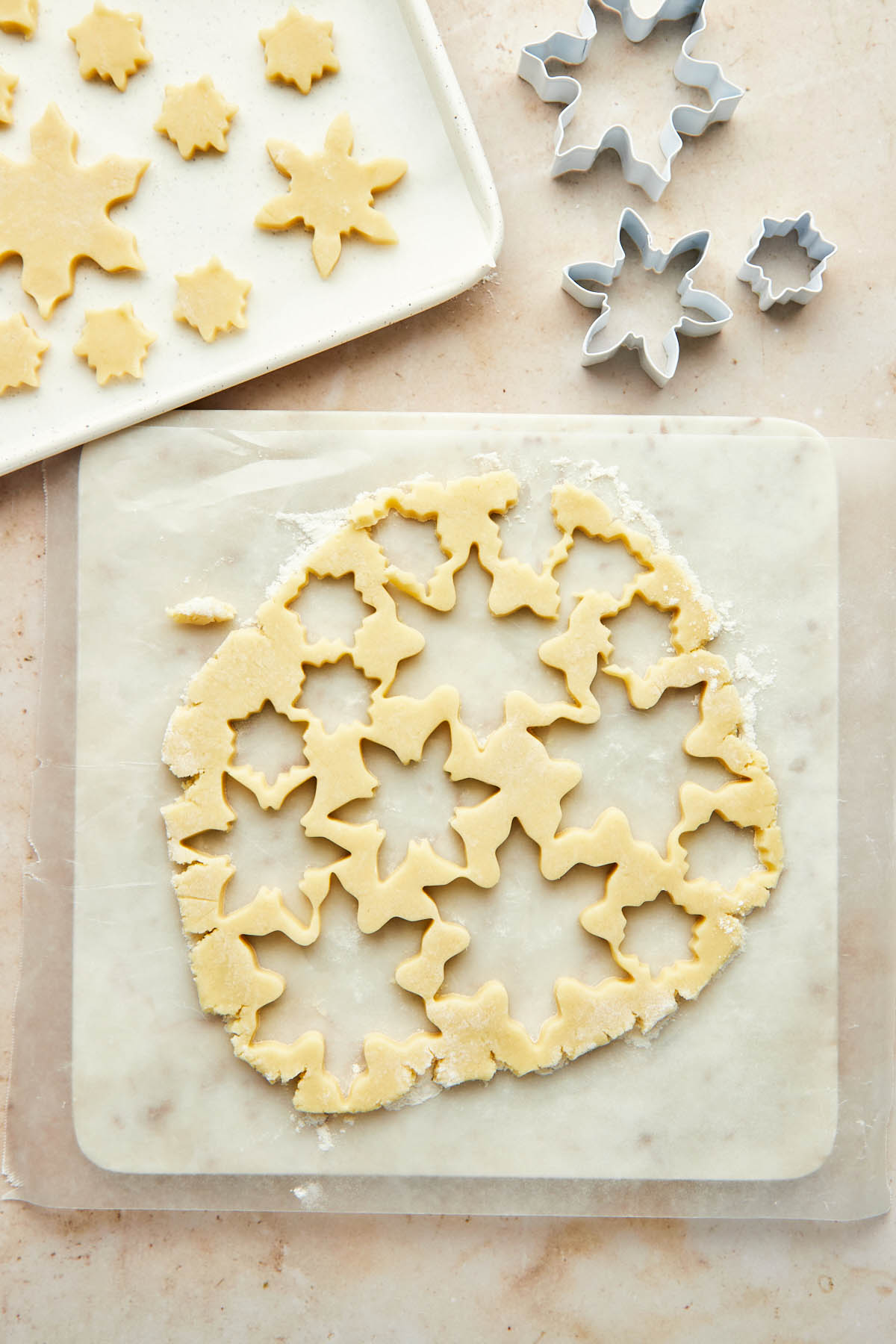 A rolled out disc of cookie dough with empty snowflake-shaped holes and a pan of snowflake-shaped cutouts nearby.