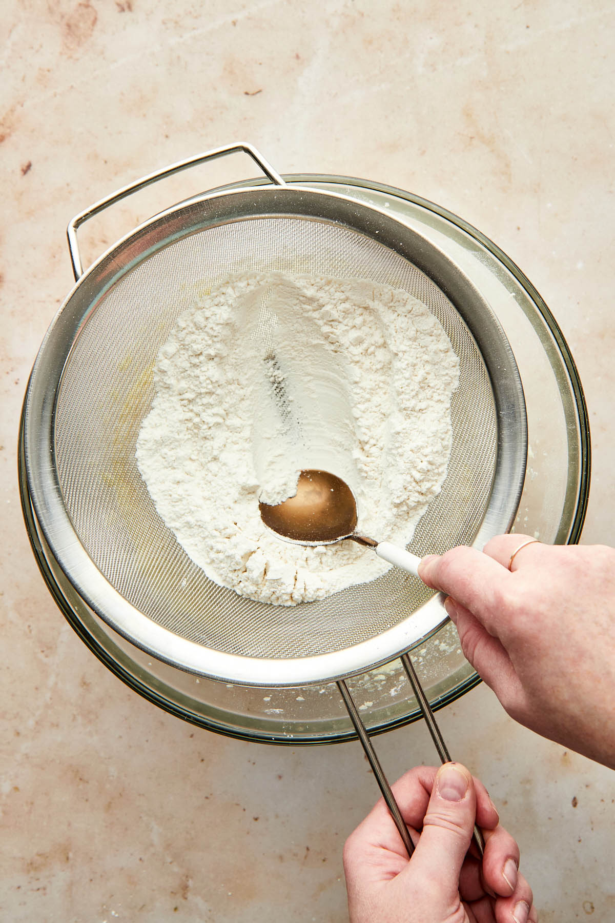 A hand using the back of a soup spoon to press flour through a sieve into a bowl of wet cookie ingredients.
