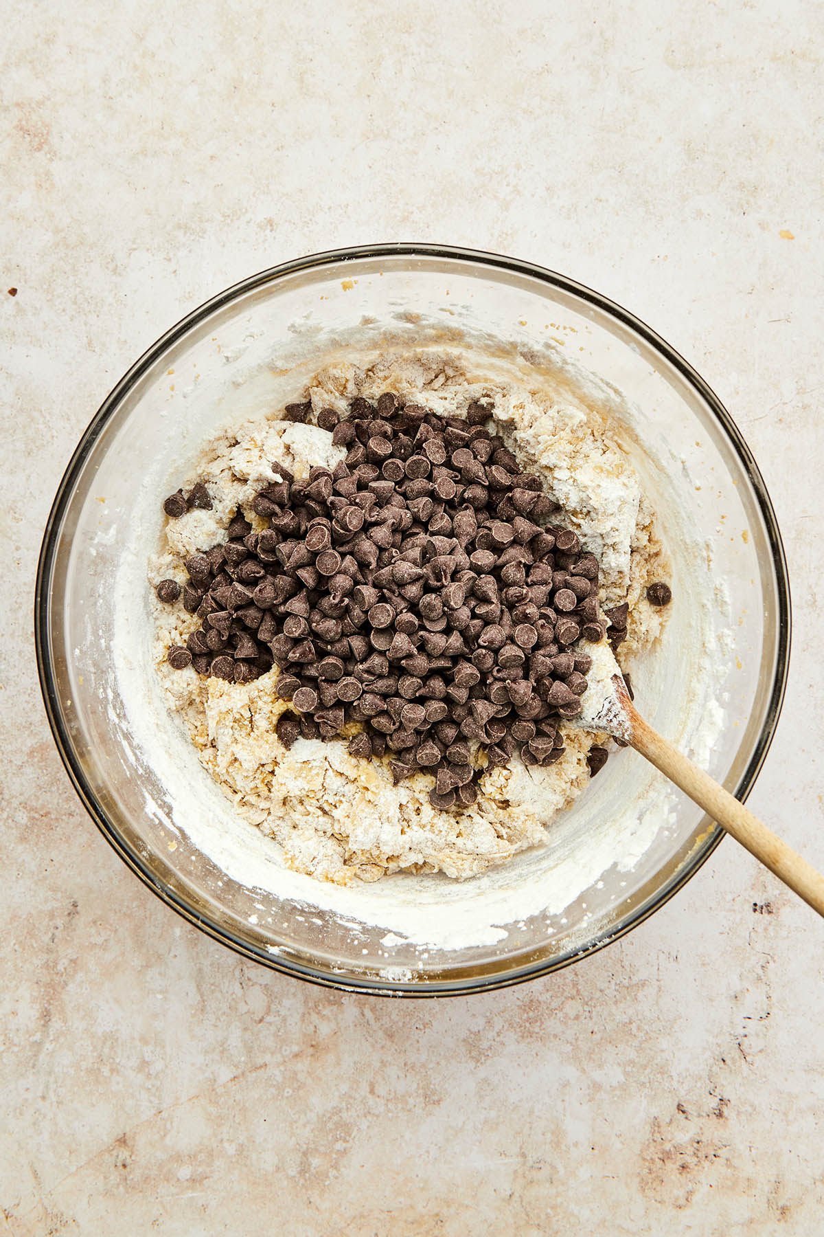 Half mixed cookie dough in a glass mixing bowl with chocolate chips and a wooden spoon on top.