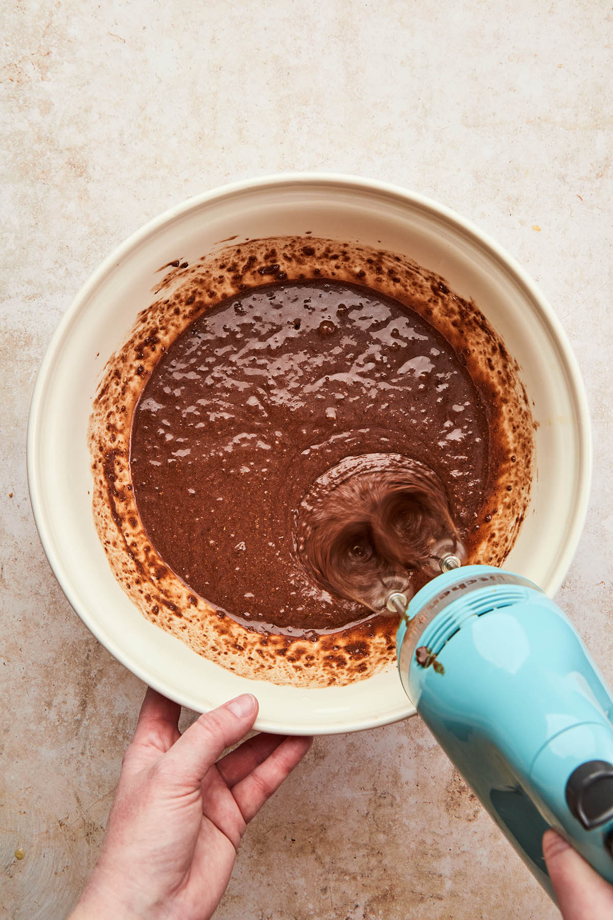 A hand using a handheld electric mixer to mix cake batter in a large mixing bowl.