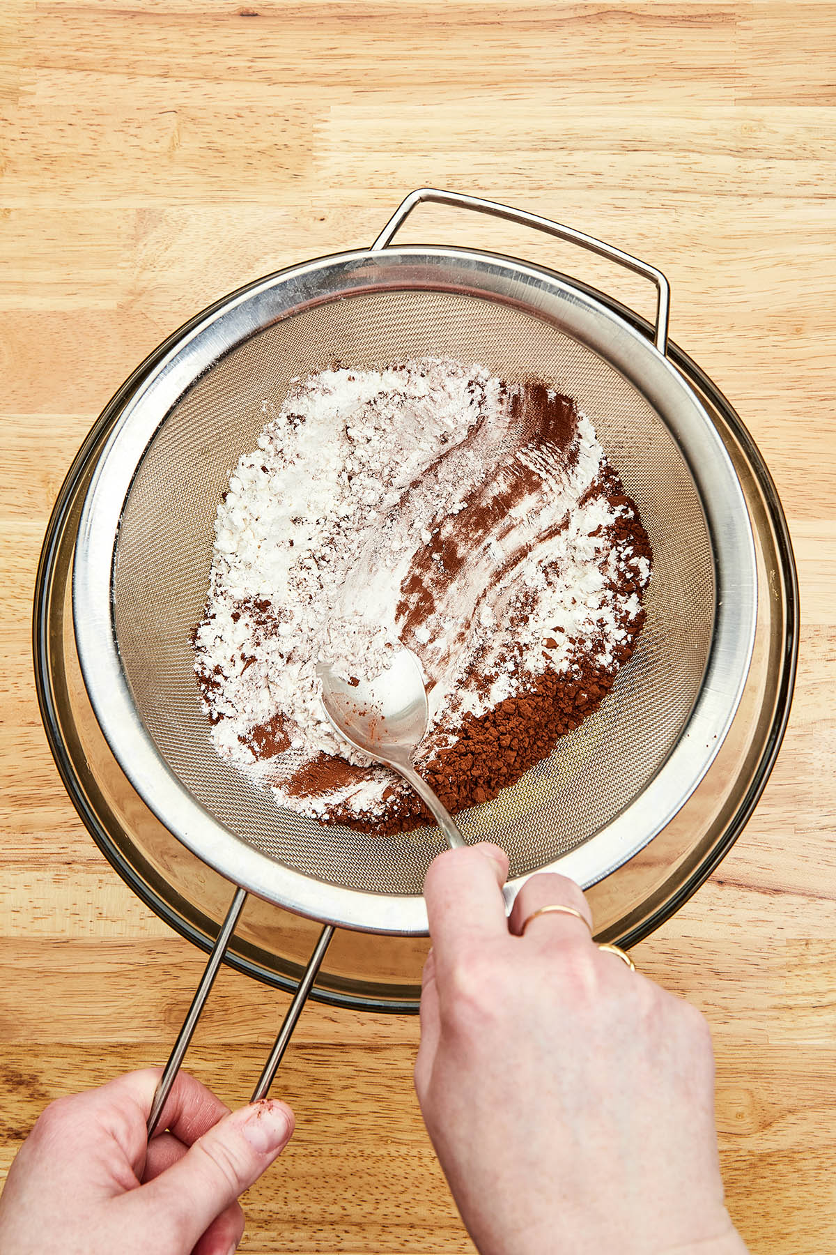A hand using the back of a spoon to press flour and cocoa through into a mixing bowl.