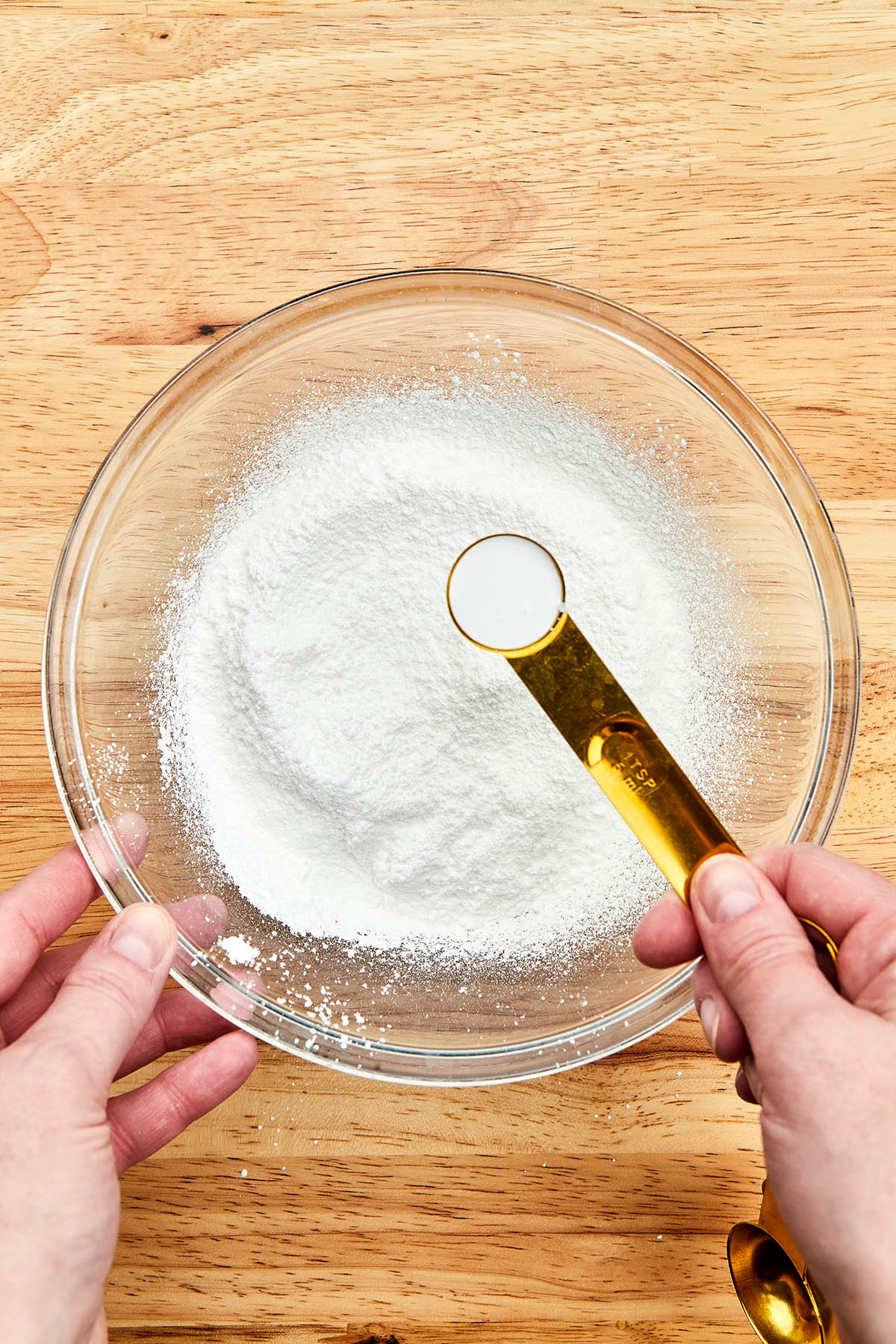 A hand holding a measuring spoon of white food colouring over a bowl of sifted powdered sugar.