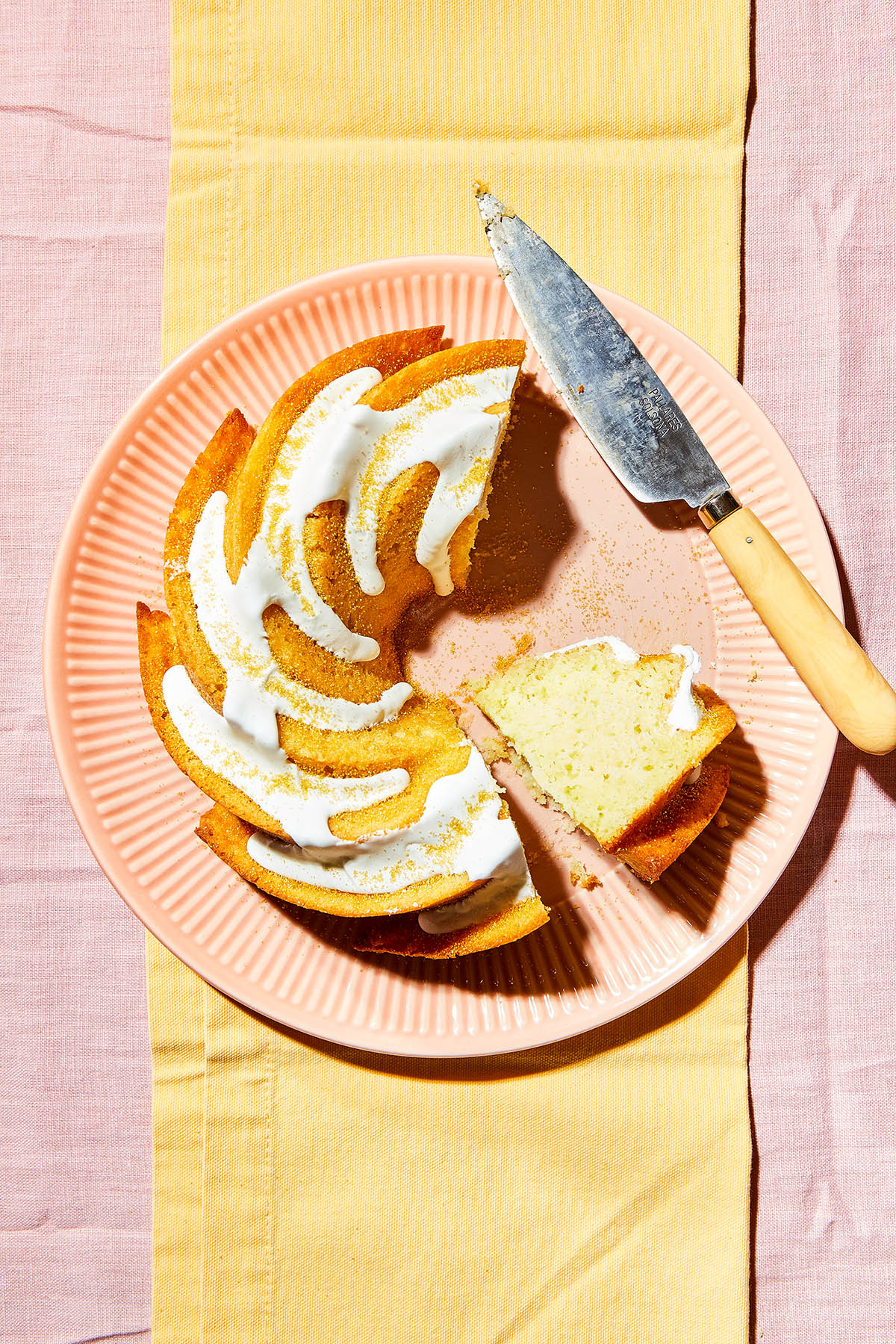 Overhead image of a lemon olive oil cake on a pink plate with one slice laying down on the plate and a knife alongside the edge of the plate.