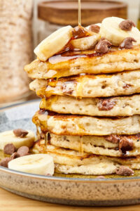 Close up image of a stack of banana chocolate chip pancakes with syrup being poured over them.