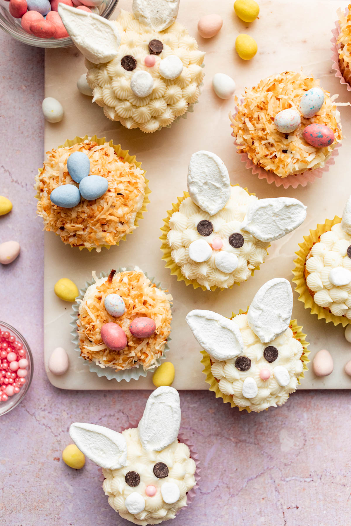 An assortment of Easter cupcakes on amarble board with chocolate eggs and sprinkles nearby.