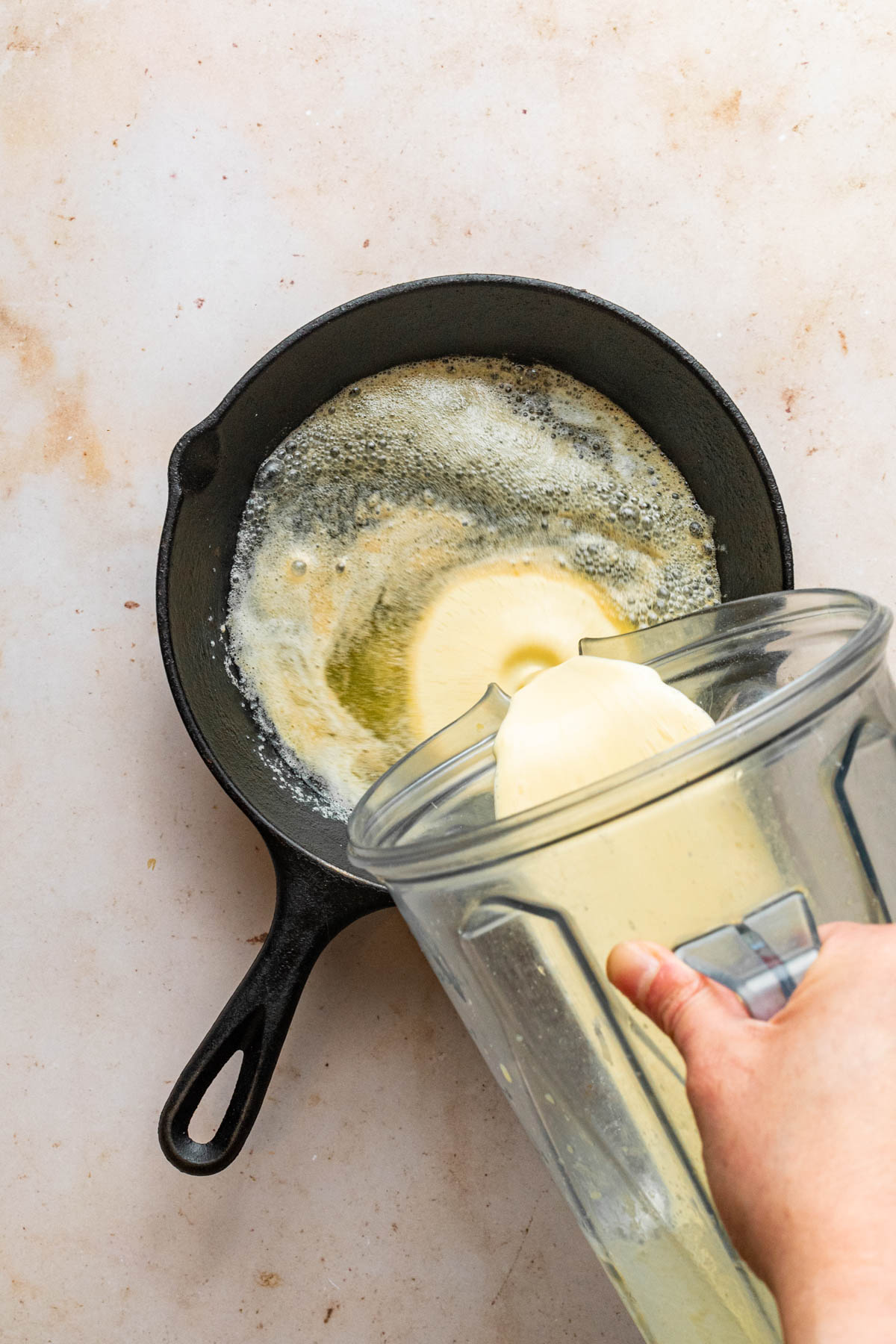 A hand pouring batter into a cast iron skillet from a blender jug.