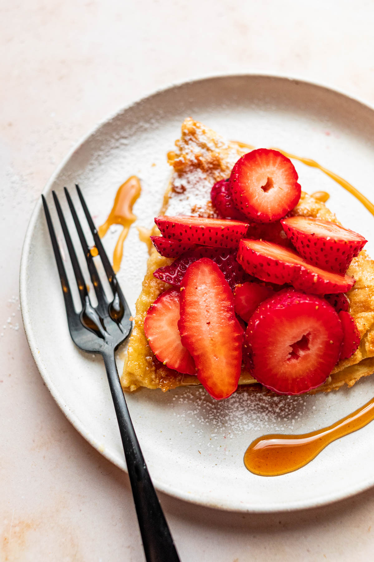 A slice of gluten-free Dutch Baby on a plate topped with strawberries, maple syrup, and powdered sugar.