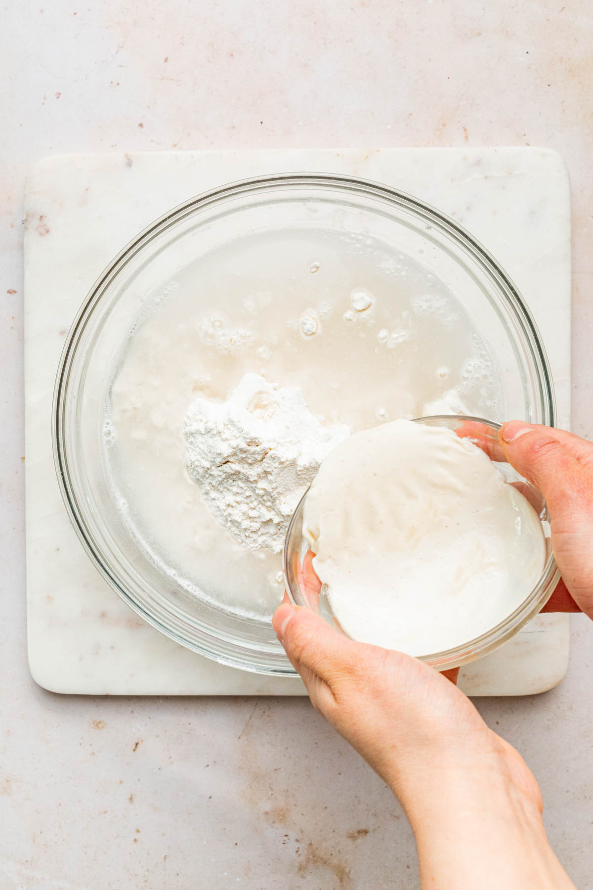 Hands holding a bowl of sourdough starter and pouring it into a bowl of water and flour.