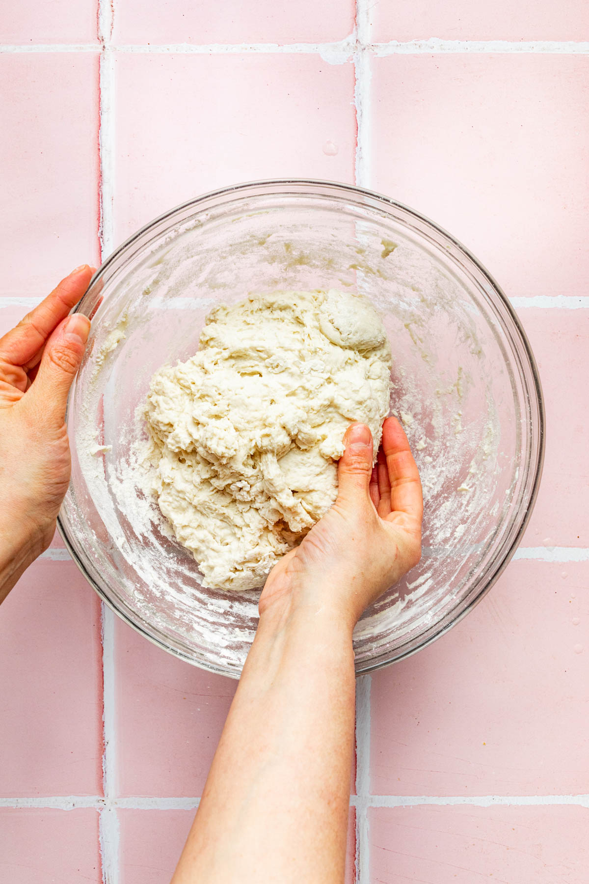 A hand reaching into a glass mixing bowl to mix shaggy dough.