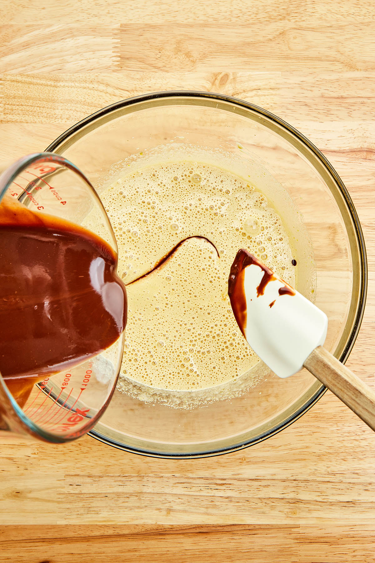 Melted chocolate being poured from a glass measuring cup into a bowl of mixed eggs and sugar.