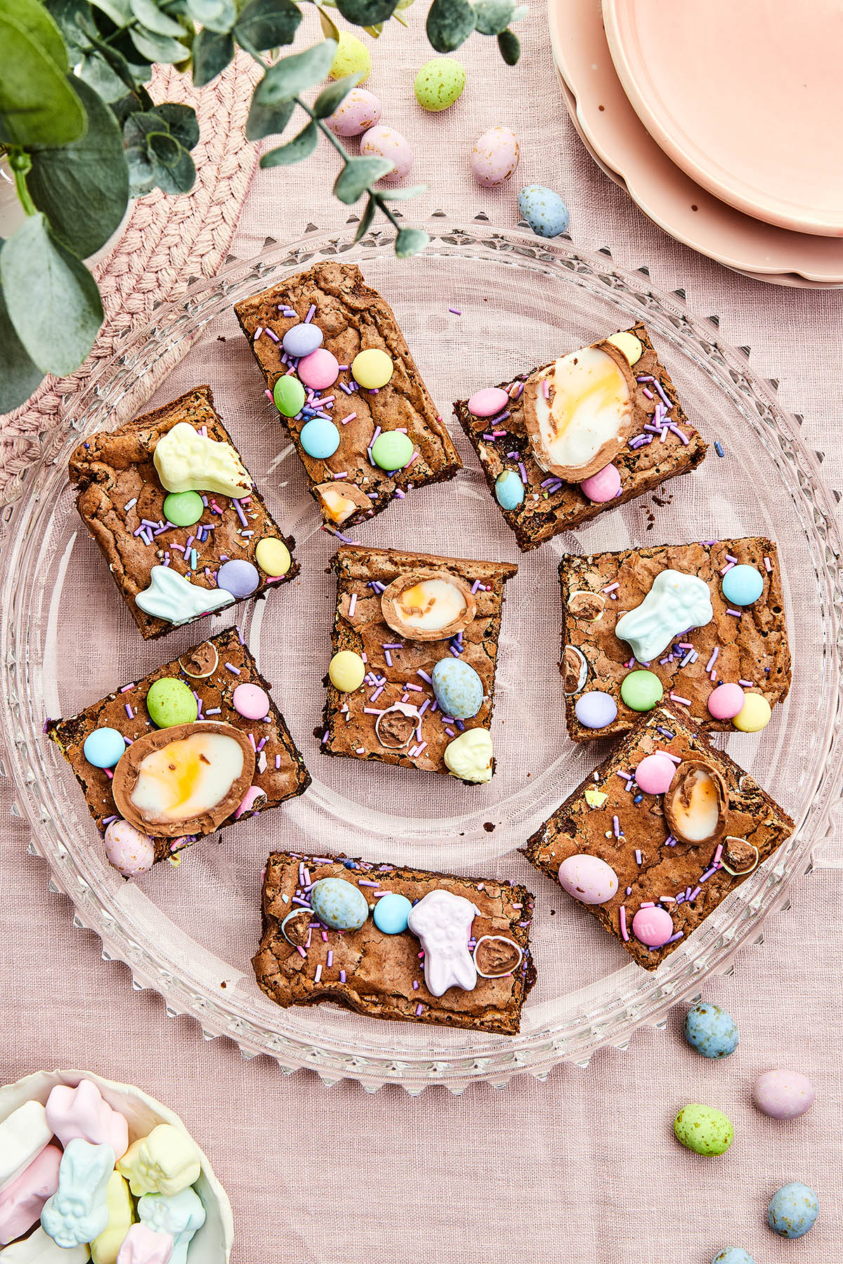 Easter brownies on a glass platter with plates, a plant, and candied chocolate eggs around.