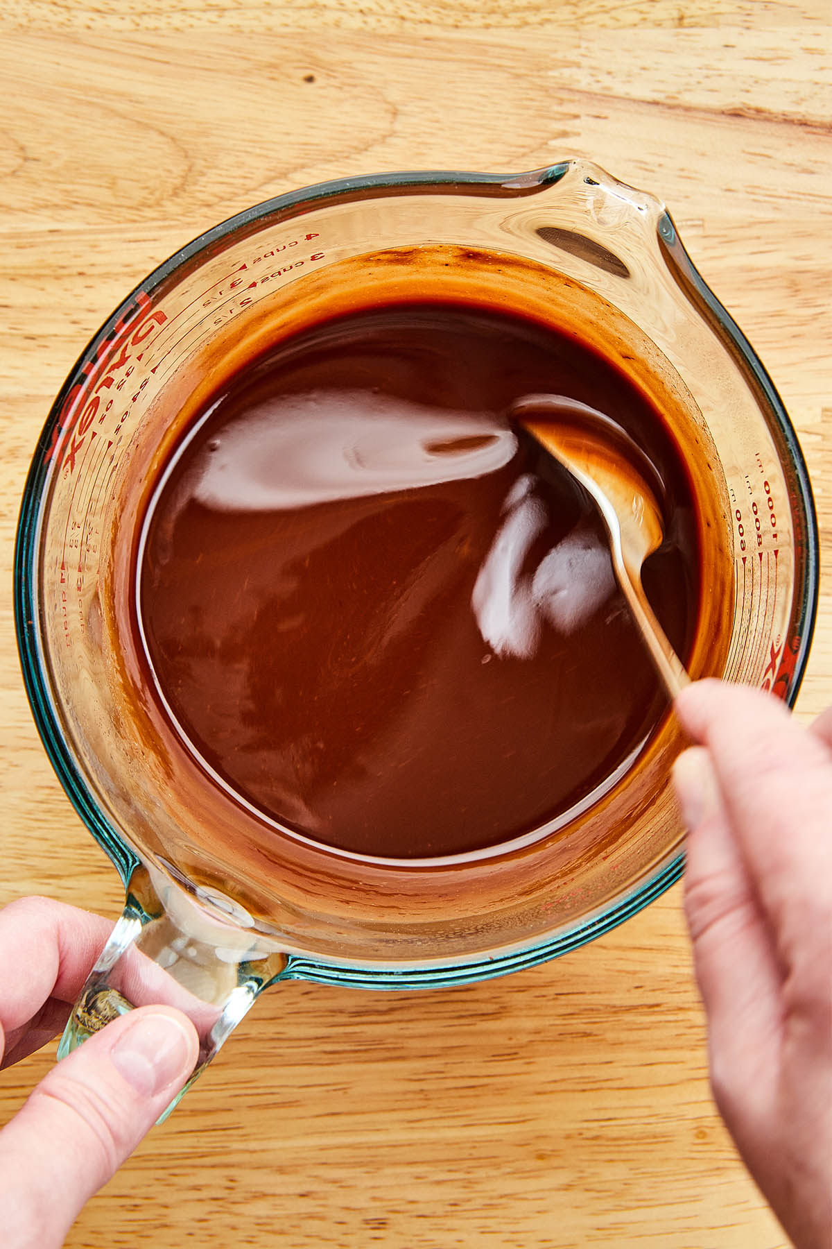 A hand using a spoon to mix melted butter and melted chocolate together in a glass measuring cup.