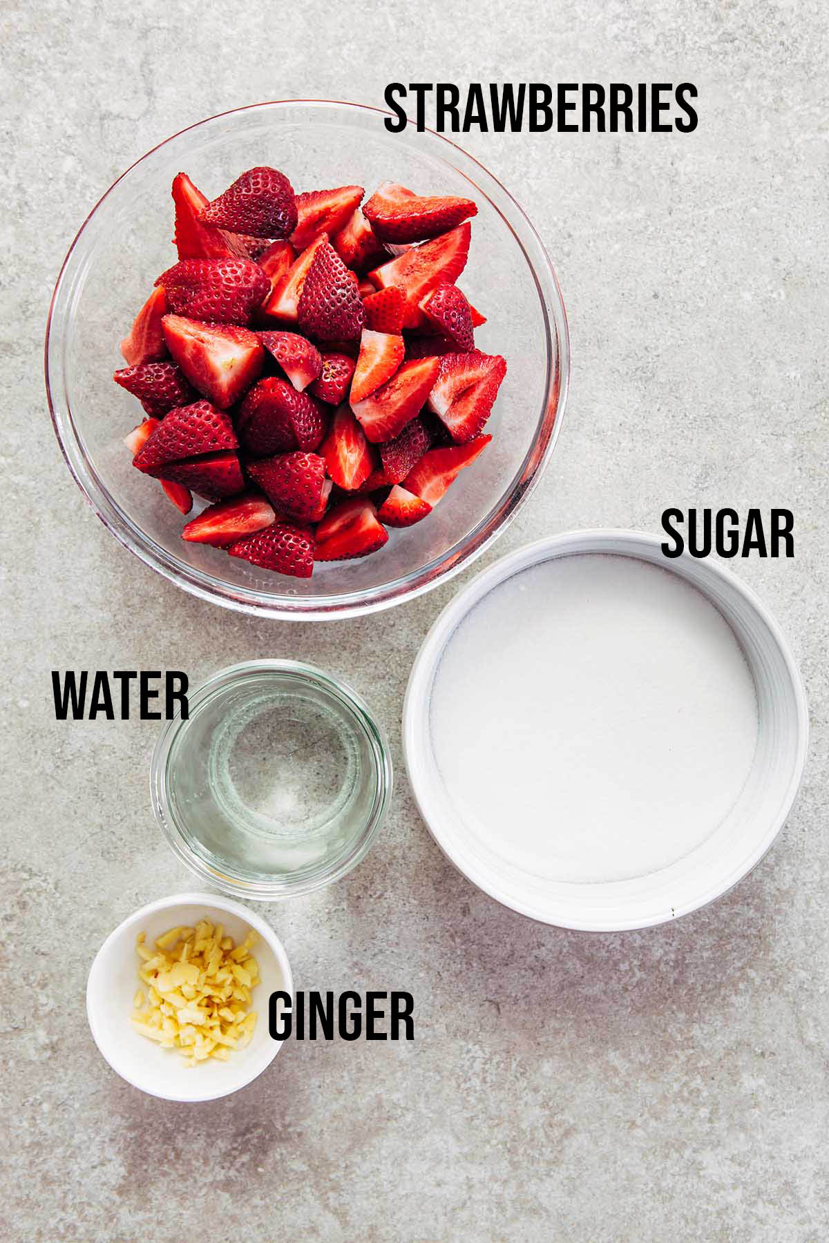 A bowl of quartered strawberries, a bowl of sugar, a jar of water, and a small bowl of minced ginger shot from overhead on a stone surface.