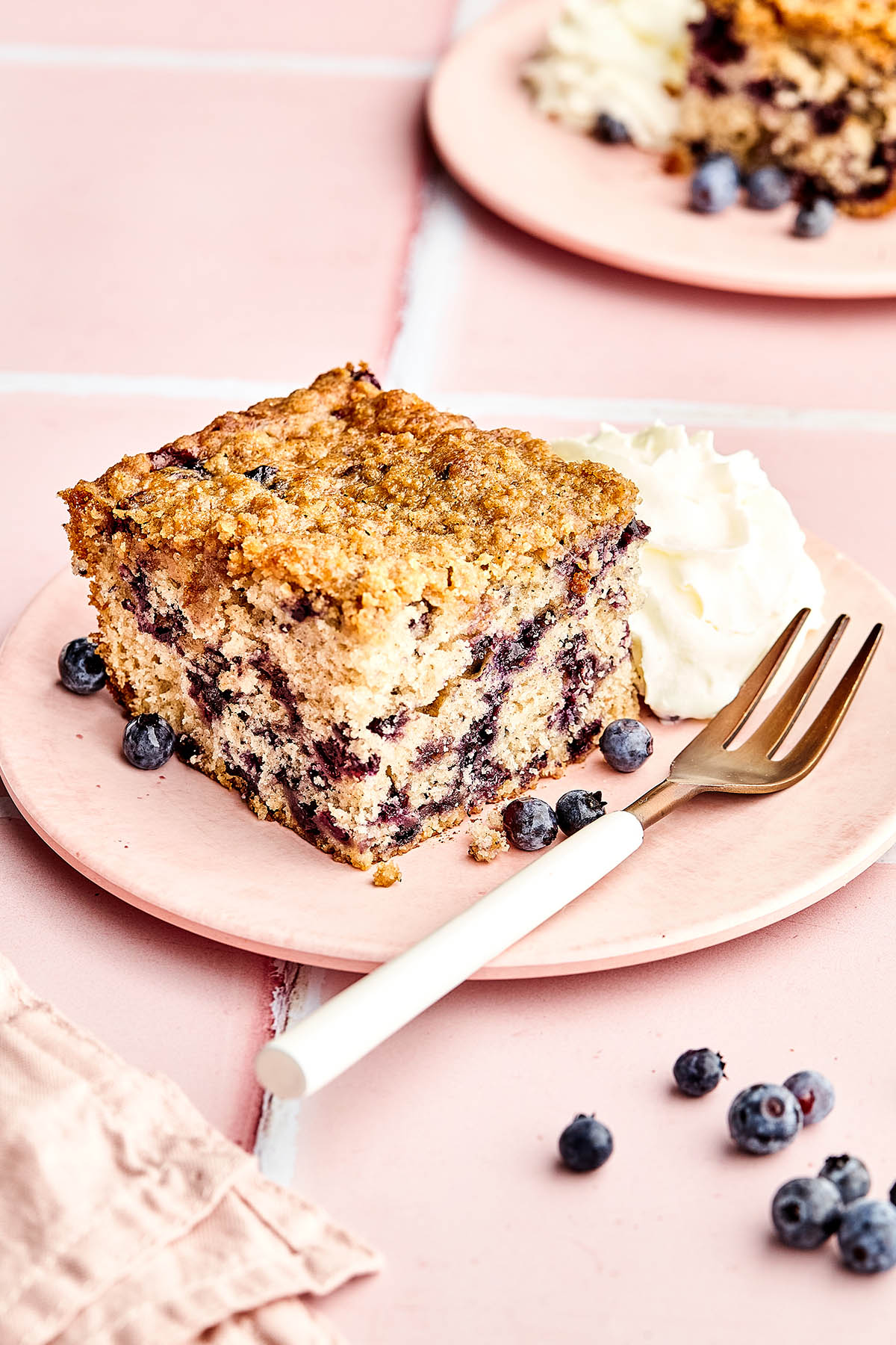 A slice of blueberry coffee cake on a pink plate with a dollop of whipped cream, a fork, and fresh blueberries scattered around.
