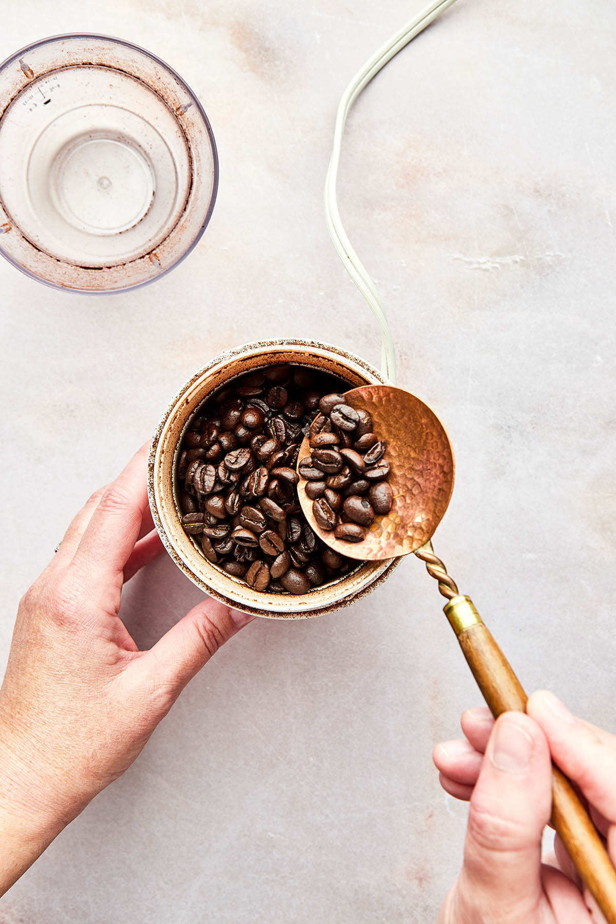 A hand using a copper spoon to portion coffee beans into a coffee grinder.
