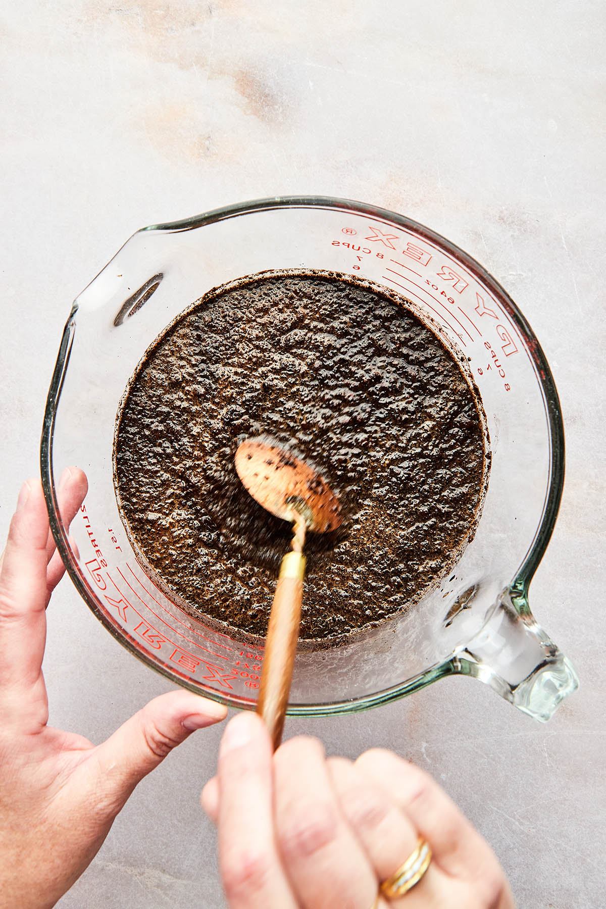 A hand using a copper spoon to stir coffee grounds and water together in a large measuring cup.