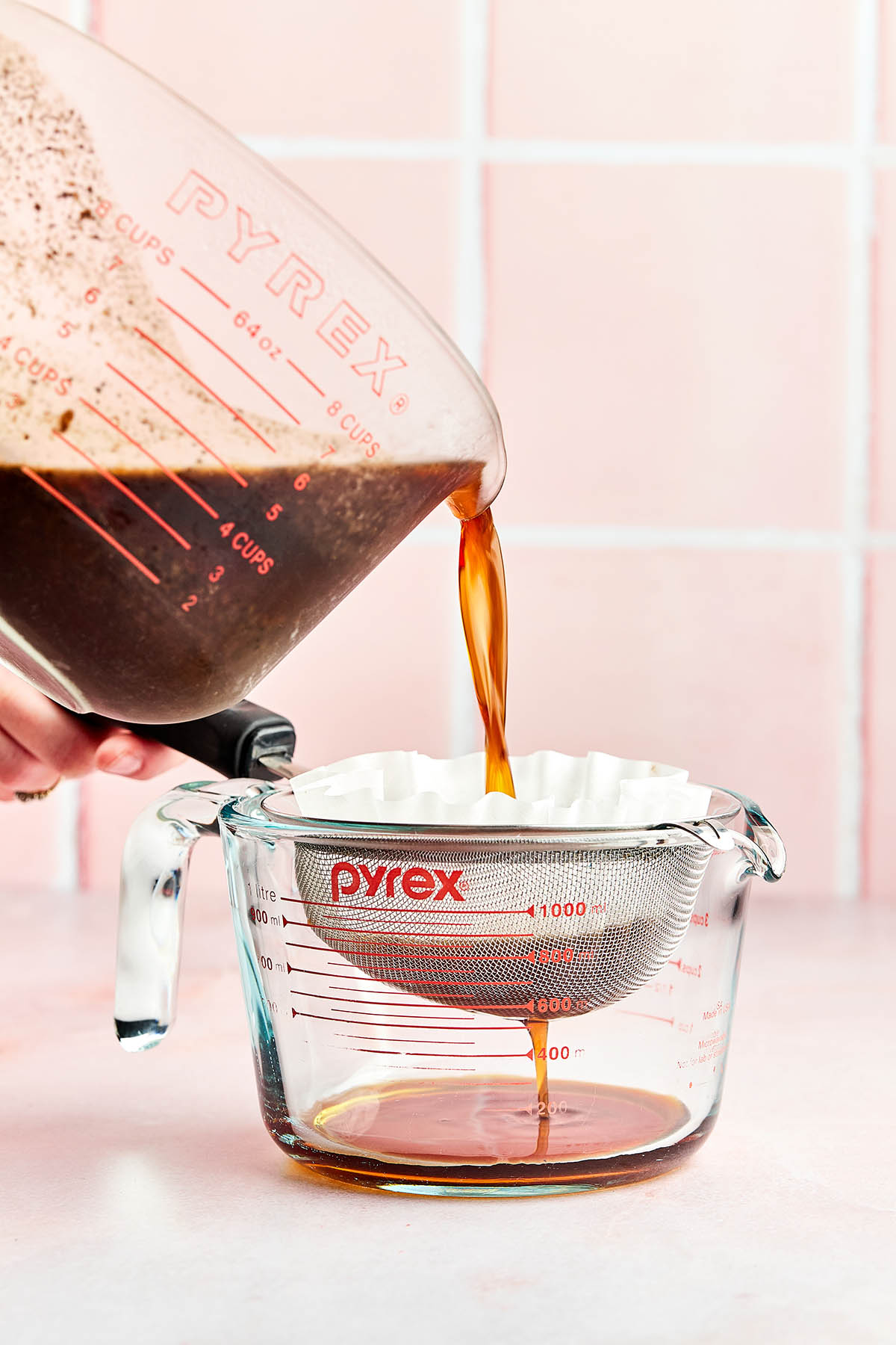 Coffee concentrate being poured from a measuring cup into another measuring cup through a fine mesh strainer lined with a coffee filter.