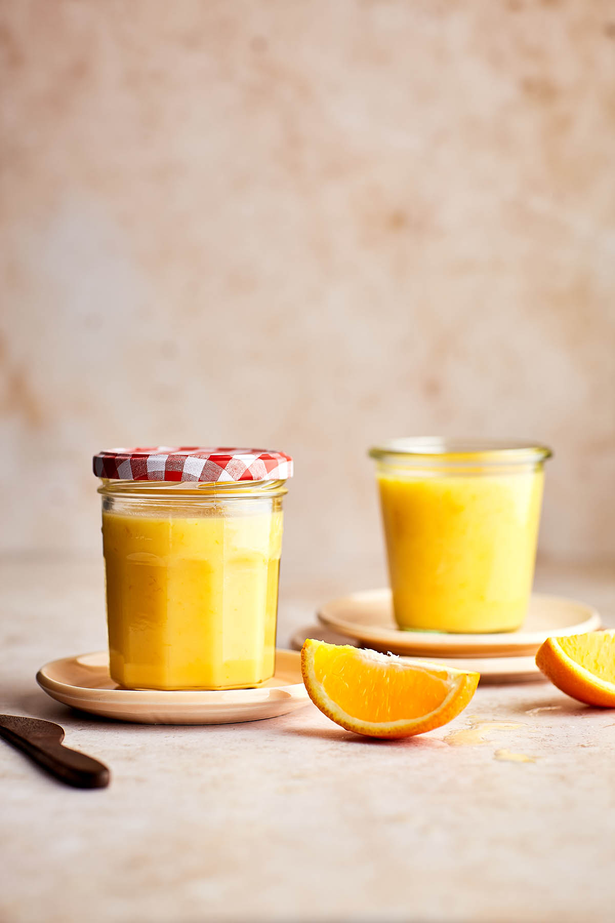 Two jars of orange curd on plates with slices of oranges nearby.